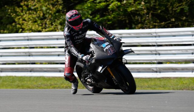 Motorcycle racer in black and red leathers leaning into a turn on a racing bike with the number 43 on the front fairing, on a track with a metal guardrail in the background.