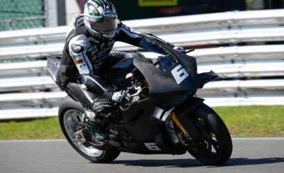 Motorcycle racer in full protective gear leaning into a turn on a race track, black sport bike with number 6 in view behind white guardrails.