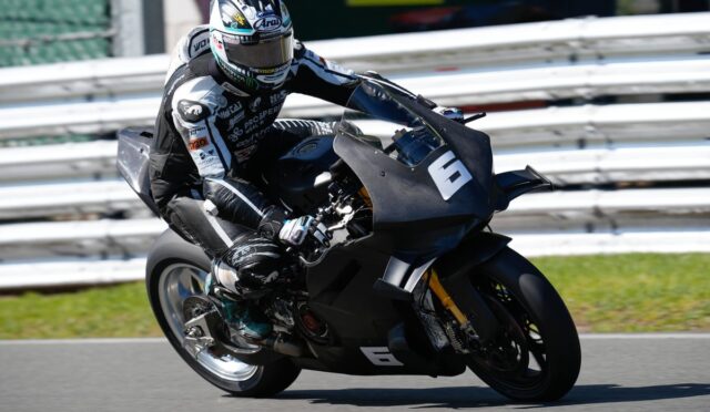 Motorcycle racer in full protective gear leaning into a turn on a race track, black sport bike with number 6 in view behind white guardrails.