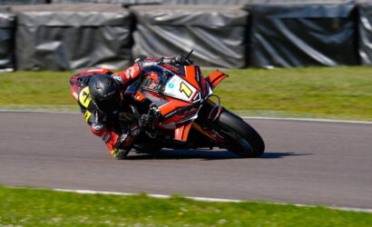 Racer on a red sports bike leaning into a sharp turn on a race track, helmeted in black gear with number plate visible.