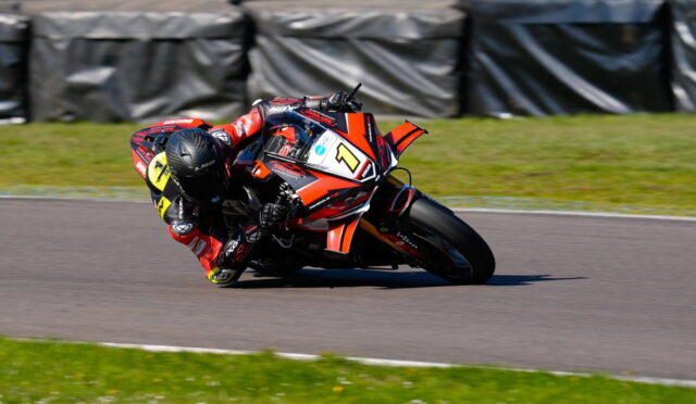 Racer on a red sports bike leaning into a sharp turn on a race track, helmeted in black gear with number plate visible.