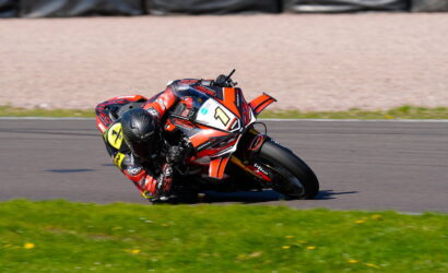 Motorcycle racer in red gear leaning into a corner on a racing track, bike numbered 1 with orange accents on a green field edge.