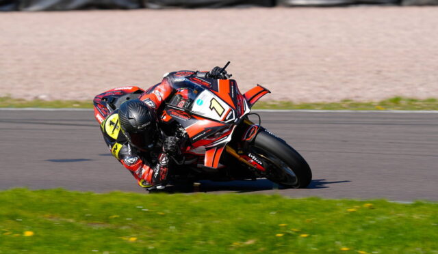 Motorcycle racer in red gear leaning into a corner on a racing track, bike numbered 1 with orange accents on a green field edge.