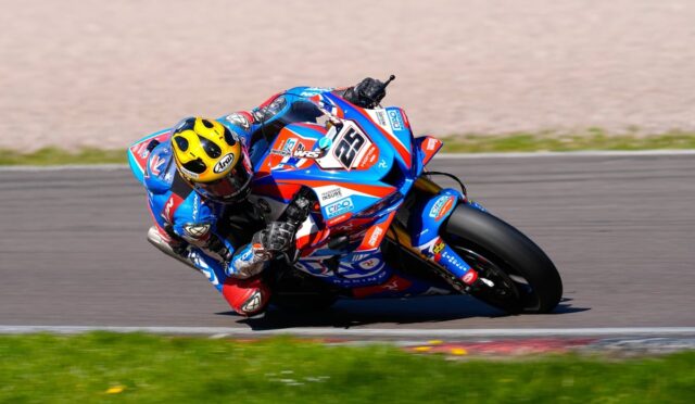 Motorcycle racer in blue-red suit leaning into a turn on a racetrack, yellow helmet visible, blue race bike number 56.](https://)