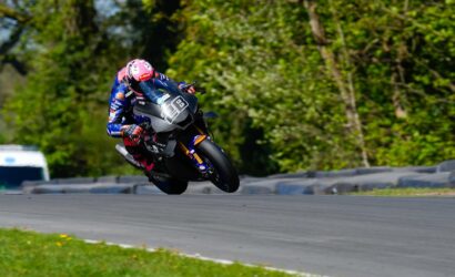 Moto race rider in blue gear and pink helmet leaning into a left-hand turn on a race track, bike number 98, trees in the background.