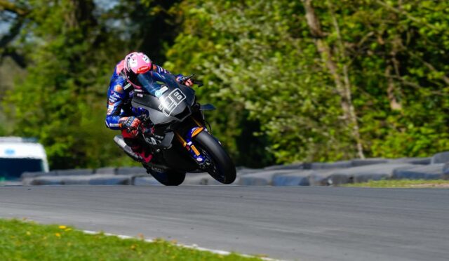 Moto race rider in blue gear and pink helmet leaning into a left-hand turn on a race track, bike number 98, trees in the background.