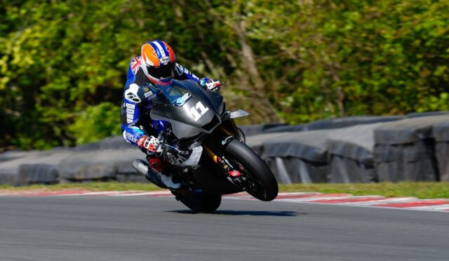 Motorcycle racer in blue gear and orange helmet leans into a turn on a race track.