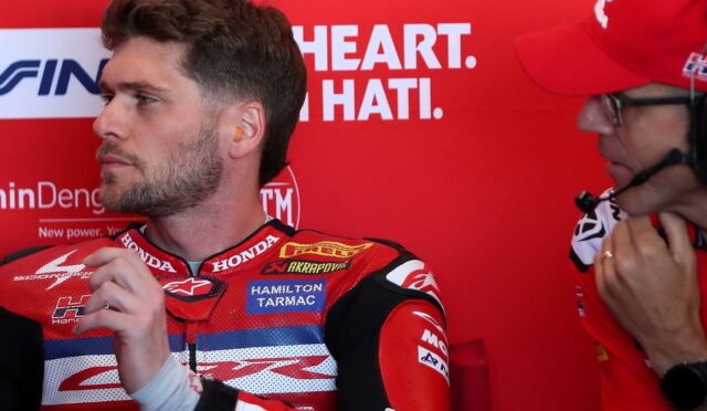 Side profile of a motorcycle racer in a red Honda racing suit, with an orange earplug visible, sitting in a pit area while a crew member in a red cap adjusts a headset nearby against a red backdrop.