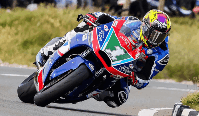 Racing motorcyclist leaning into a right-hand curve on a blue-and-red sport bike in full gear with a neon helmet.
