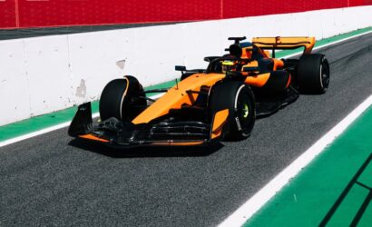 Orange Formula 1 car on a race track near a white barrier with red wall above, green run-off on the side.