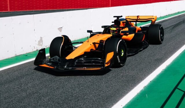 Orange Formula 1 car on a race track near a white barrier with red wall above, green run-off on the side.