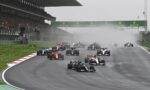 Formula 1 race start on a wet track with spray, under a Rolex banner and packed grandstands in the background.