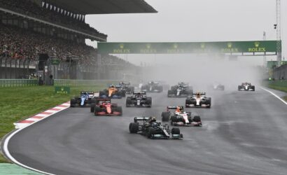 Formula 1 race start on a wet track with spray, under a Rolex banner and packed grandstands in the background.