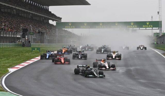 Formula 1 race start on a wet track with spray, under a Rolex banner and packed grandstands in the background.