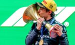 F1 driver in a Mercedes suit kisses a large gold trophy on a bright green backdrop, medal around neck