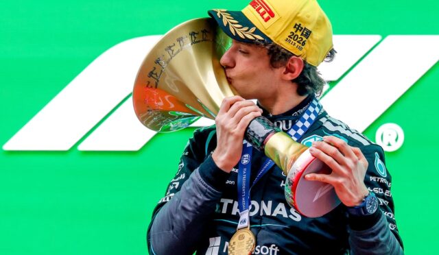 F1 driver in a Mercedes suit kisses a large gold trophy on a bright green backdrop, medal around neck
