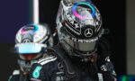 Close-up of a Formula 1 driver in a black Mercedes helmet and suit, with Petronas logos, standing in the pit area (second driver blurred in background).