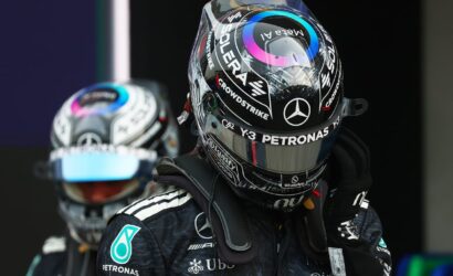 Close-up of a Formula 1 driver in a black Mercedes helmet and suit, with Petronas logos, standing in the pit area (second driver blurred in background).