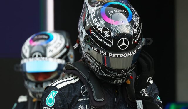 Close-up of a Formula 1 driver in a black Mercedes helmet and suit, with Petronas logos, standing in the pit area (second driver blurred in background).
