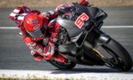 MotoGP rider in a red racing suit and red helmet leans into a sharp turn on a Ducati race bike on a race track, sponsor logos visible on suit and bike.