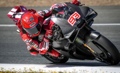 MotoGP rider in a red racing suit and red helmet leans into a sharp turn on a Ducati race bike on a race track, sponsor logos visible on suit and bike.