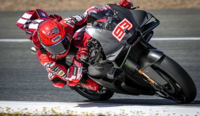 MotoGP rider in a red racing suit and red helmet leans into a sharp turn on a Ducati race bike on a race track, sponsor logos visible on suit and bike.