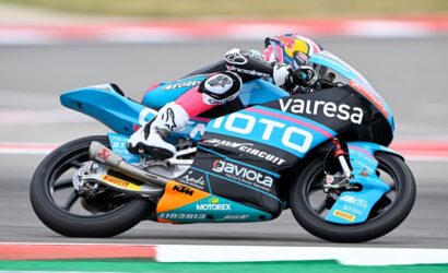 Motorcycle racer leaning into a high-speed turn on a blue race bike with sponsor logos.