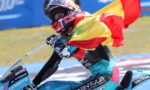 Racer on a blue motorcycle waves a Spanish flag during a victory lap, helmeted and in colorful racing gear.