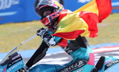 Racer on a blue motorcycle waves a Spanish flag during a victory lap, helmeted and in colorful racing gear.