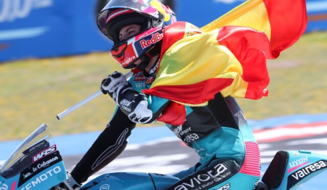 Racer on a blue motorcycle waves a Spanish flag during a victory lap, helmeted and in colorful racing gear.