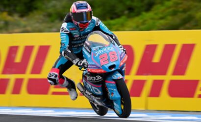 Motorcycle racer in blue suit and pink helmet leaping a curb on a race bike number 28, blue livery, DHL barriers in background.
