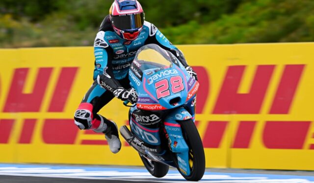 Motorcycle racer in blue suit and pink helmet leaping a curb on a race bike number 28, blue livery, DHL barriers in background.
