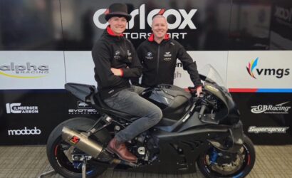 Two men posing on a matte black sport motorcycle in a showroom, wearing black jackets with sponsor logos and a backdrop of racing banners behind them.