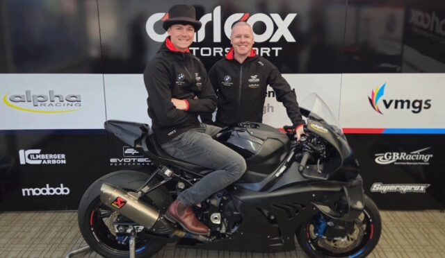 Two men posing on a matte black sport motorcycle in a showroom, wearing black jackets with sponsor logos and a backdrop of racing banners behind them.