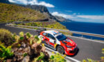 Red rally car navigating a winding coastal mountain road with blue sea in the background and guardrails nearby.