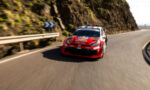 Red rally car speeding on a winding coastal road with a guardrail and rocky cliff on the left.