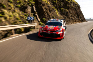 Red rally car speeding on a winding coastal road with a guardrail and rocky cliff on the left.