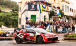 Red Bull rally car speeding through a street rally as crowds watch from behind barriers.