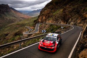 Red rally car speeding around a winding mountain road with guardrails and canyon in the background.