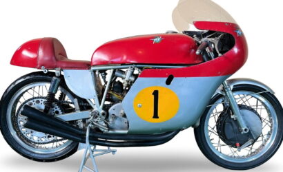Vintage racing motorcycle with red top, blue side panel, and a yellow number 1 decal on the side panel, on a stand.