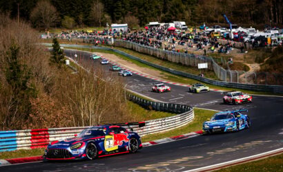 Race cars power through a tight turn on a race track with a large crowd watching from the grassy spectator area nearby.