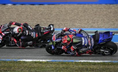 Two professional motorcycle racers lean into a turn on a race track, blue Yamaha bike in front with Monster energy branding and black/red Aprilia bike behind.