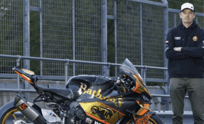 Man in a navy hoodie and cap stands with arms crossed beside a black and gold sport motorcycle by a metal fence at a racetrack area.
