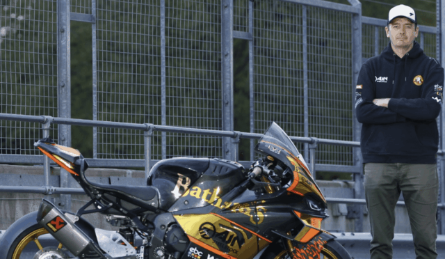 Man in a navy hoodie and cap stands with arms crossed beside a black and gold sport motorcycle by a metal fence at a racetrack area.