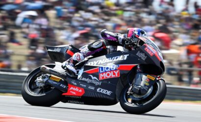 Motorcycle racer leaning into a turn on a sponsor-laden racing bike at a track with blurred crowd in background