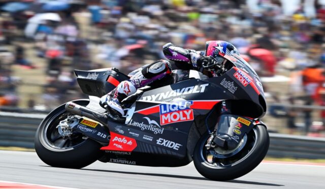 Motorcycle racer leaning into a turn on a sponsor-laden racing bike at a track with blurred crowd in background