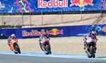 Three motorcycle racers lean into a curve on a track with Red Bull banners in the background.