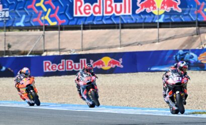 Three motorcycle racers lean into a curve on a track with Red Bull banners in the background.