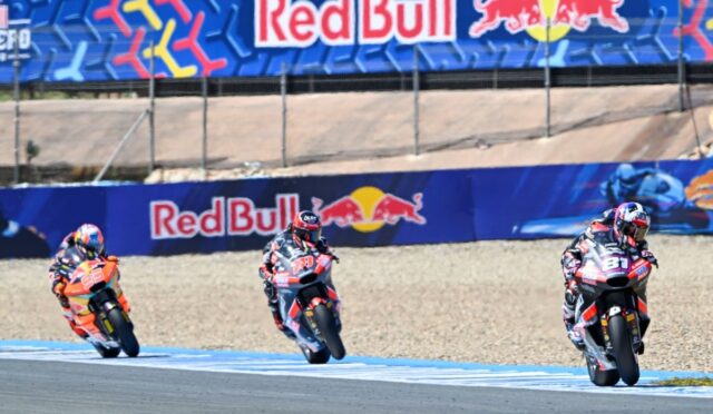 Three motorcycle racers lean into a curve on a track with Red Bull banners in the background.