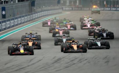 Group of colorful Formula 1 cars racing side by side on a track, near the start of the race with barriers along the left.